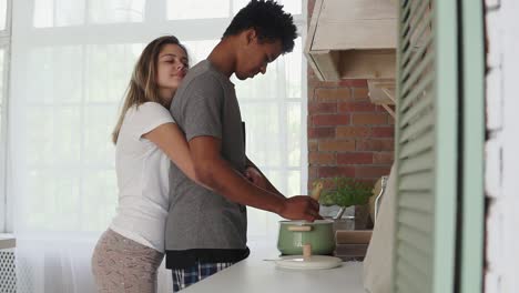 hombre africano preparando comida en una olla de pie en la cocina. su novia caucásica se le acerca y lo abraza. amor verdadero