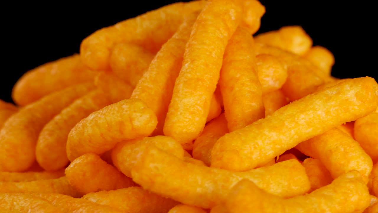 Golden cheese puffs rotate in macro view under studio lighting against a clean black background