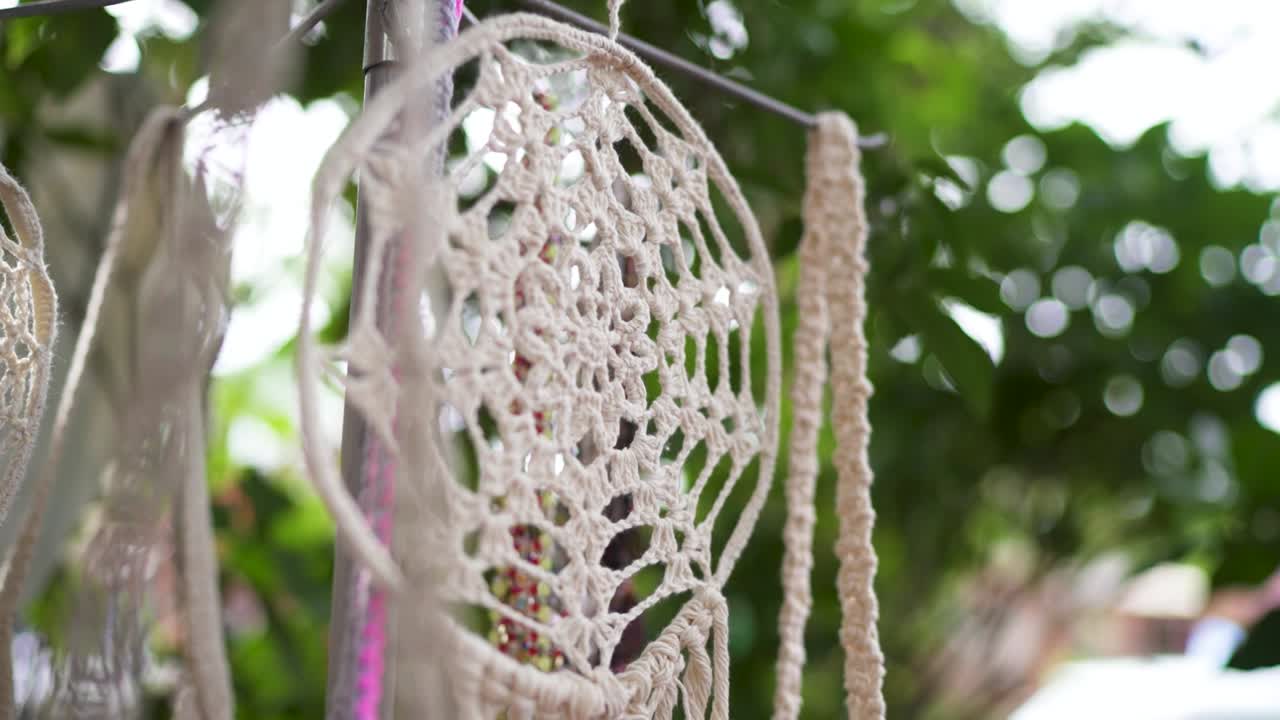A beautiful close-up of a handmade dreamcatcher, intricately woven in a crochet style. It hangs outdoors, gently moving, with a soft, out-of-focus background of green foliage