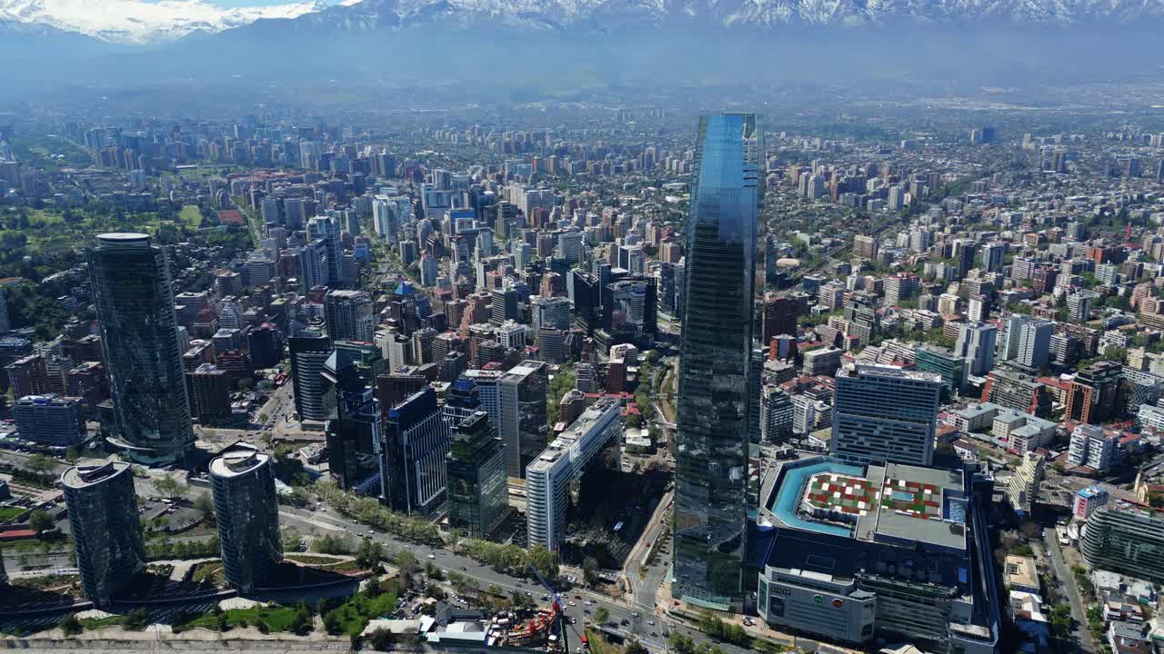 Drone aerial orbit revealing Costanera skyscrapers, highways and snowcapped Andes under clear daytime skies. Santiago, Chile