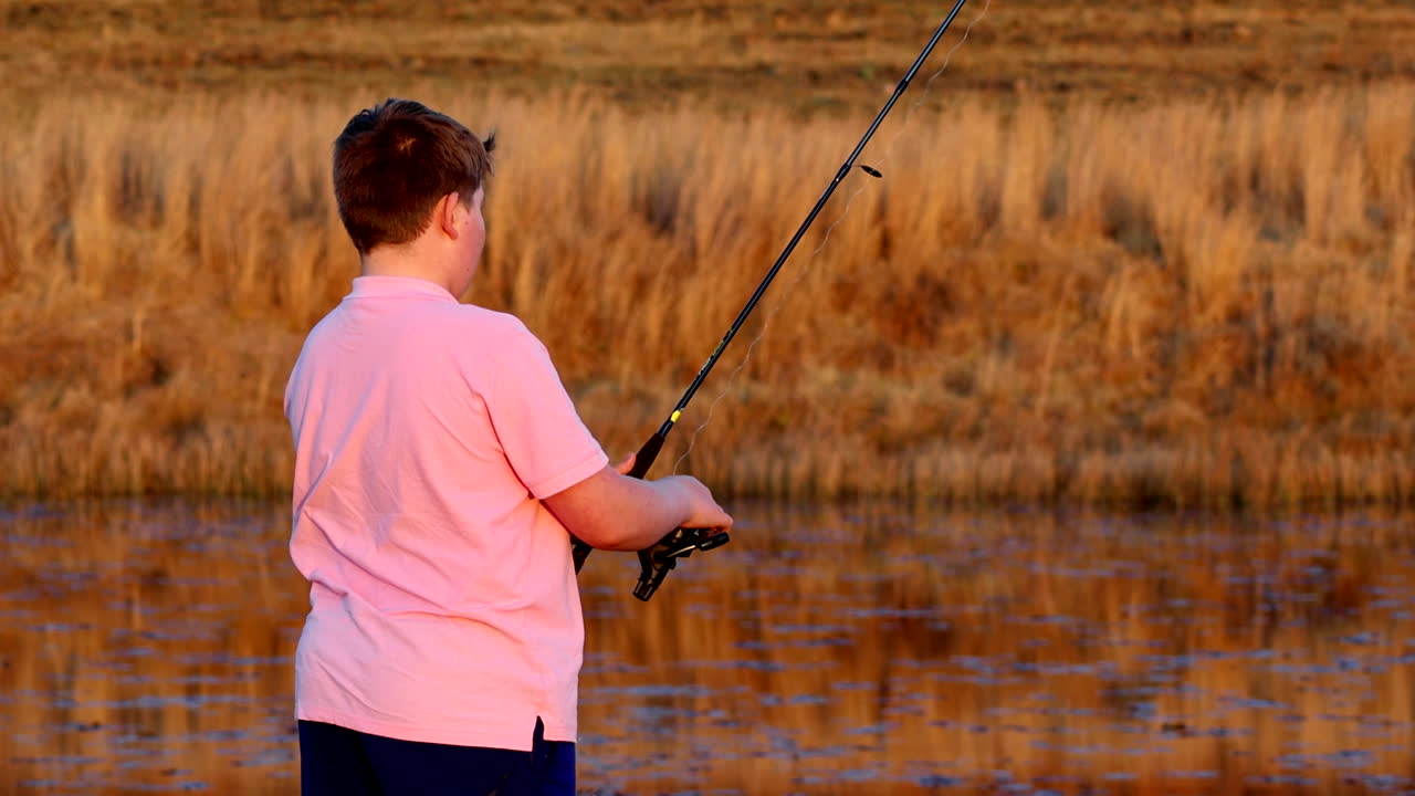 젊은이가 평온한 호수에 시줄을 던지고, 밝은 분홍색 티셔츠를 입은 백인 남자가 자연의 평화와 조용함을 즐기고 있습니다.