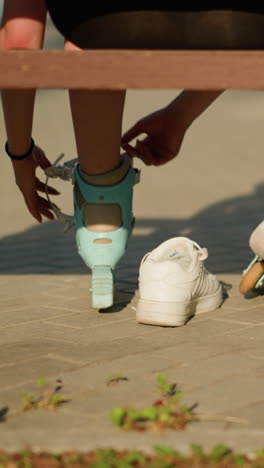 back view of woman strapping roller skate on right leg while other roller skate rests on left leg, with left leg slightly out of sneaker, seated on bench outdoors, with warm sunlight in background