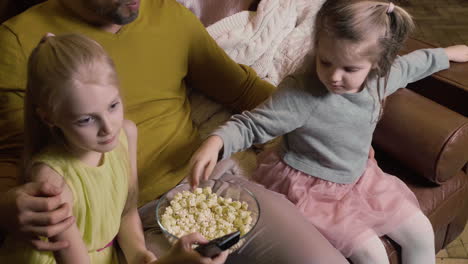 dad and his two little daughters eating popcorn and watching a movie while sitting on sofa at night at home 3