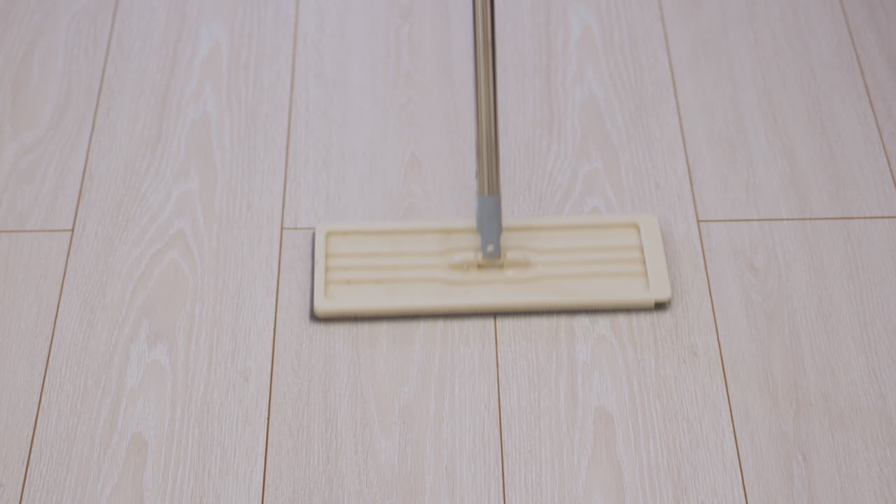 Close up of mop being pushed across tiled wooden floor indoors with person in background showing household cleaning process focused on hygiene maintenance