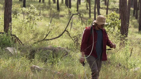 disparo medio de un guardabosques caminando con un rociador de mochila en el bosque, lleva una camisa roja y un sombrero