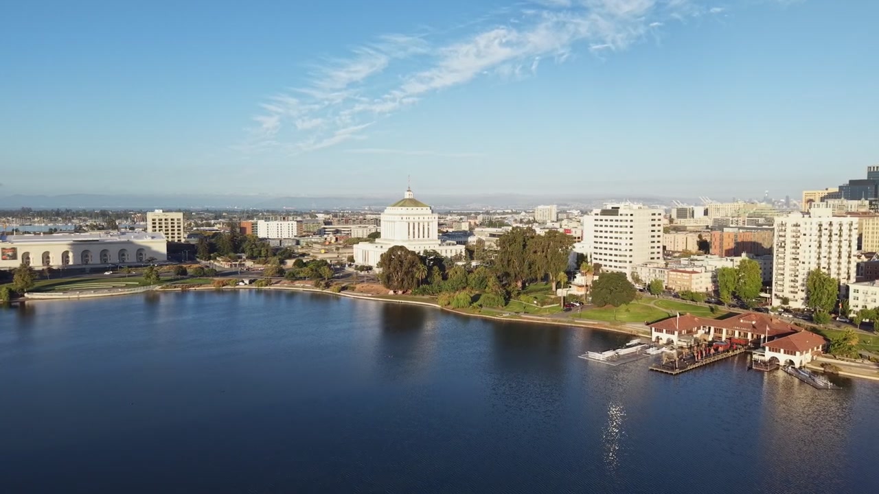 Riprese aeree di una mattina soleggiata che sorvolano il Lago Merritt, mostrando lo skyline del centro di Oakland con i suoi edifici e il parco sul lungomare sotto un cielo azzurro e limpido in una splendida giornata in California