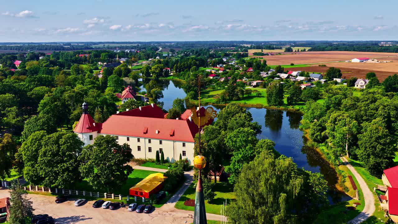 Foto aérea do Castelo de Jaunpils cercado por lagoas, árvores e terras agrícolas, mostrando a herança rural da Letônia e a vibrante paisagem de verão