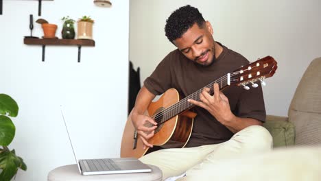 cheerful ethnic man playing guitar with laptop on sofa