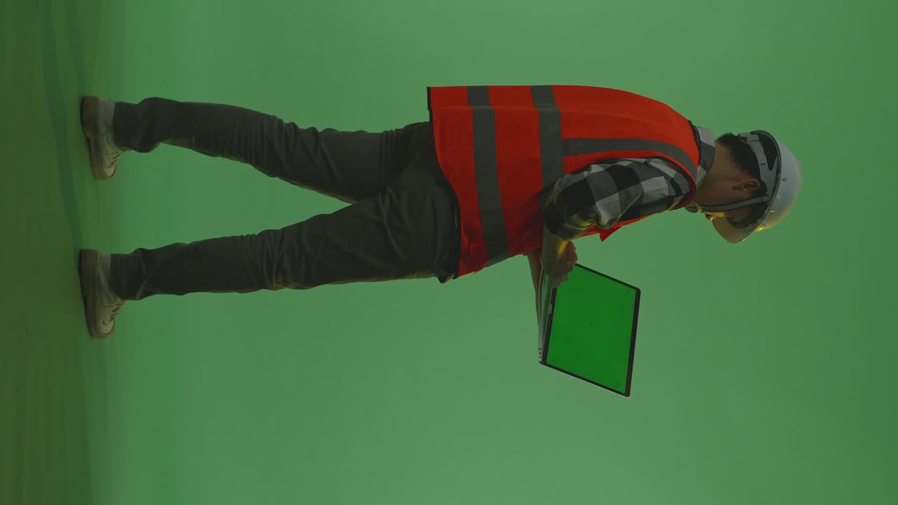 vista trasera completa del cuerpo de un ingeniero masculino asiático con casco de seguridad trabajando en una computadora portátil de pantalla verde y mirando a su alrededor mientras está de pie en el estudio de fondo de pantalla verde, durante la puesta o el amanecer