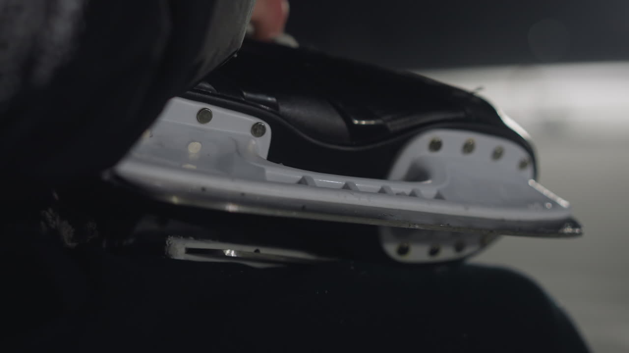 close up of individual holding black skate boot with sharp metal blade, positioned above blurred ice rink under night lighting, cold atmosphere with reflective icy surface and soft ambient glow