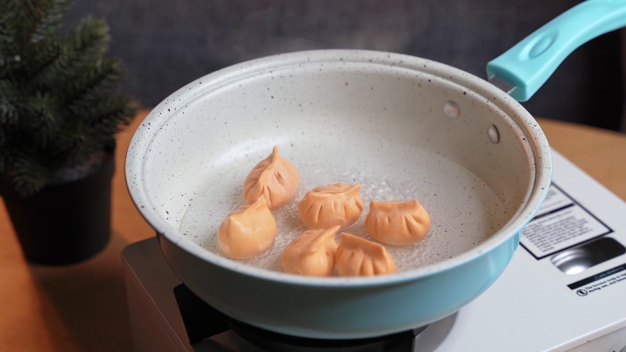 Close up shot of a person's hand placing a fresh orange dumpling into a light blue pan of boiling water. The asian dim sum cooks on a white portable stove on a wooden table indoors.