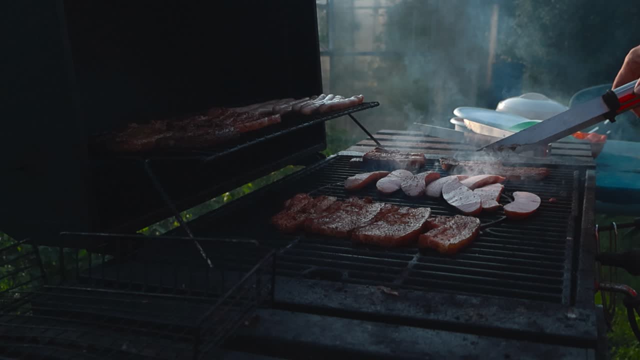 Close up view of a white male hand moving grilled and cooking meat and sausages on a smoking hot grill in a sunny garden during summer or autumn time. The meat is seasoned and has char marks on it