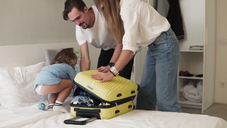 Young-caucasian-couple-tries-to-zip-suitcase-full-of-clothes.-Parents-closing-suitcase-and-ask-their-little-daughter-to-sit-on