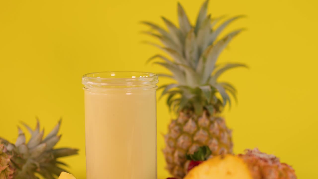 Hand adds whipped cream to pineapple smoothie with fresh fruit on vibrant yellow background