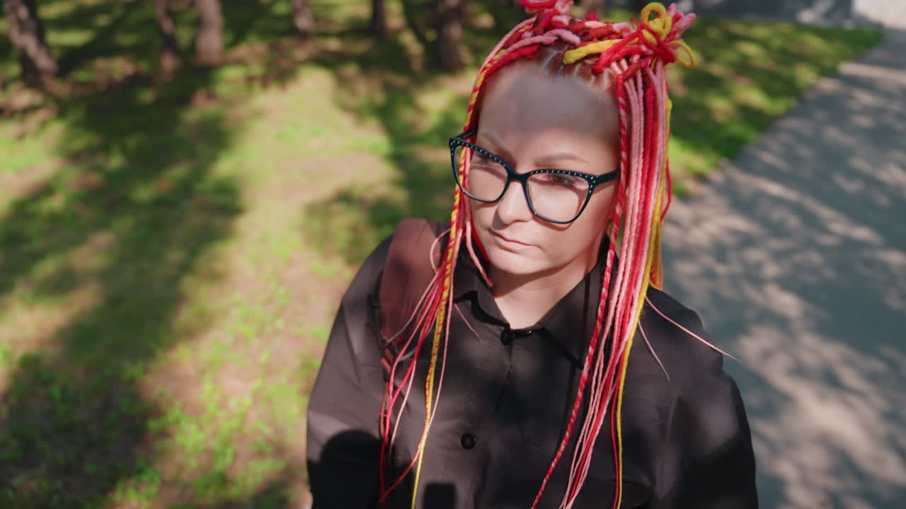 mujer blanca triste caminando por un parque, rastas de color rojo y amarillo brillante, gafas extragrandes, abrigo negro, mirada hacia abajo, paso solitario por un sendero iluminado por el sol enmarcado por árboles, estado de ánimo introspectivo