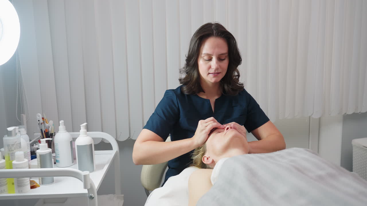 Beauty expert gently performs soothing fingertip rub on relaxed client face under blanket beside trolley with skin care tools and illuminated by ring light in serene spa treatment room