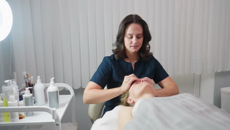 beauty expert gently performs soothing fingertip rub on relaxed client face under blanket beside trolley with skin care tools and illuminated by ring light in serene spa treatment room