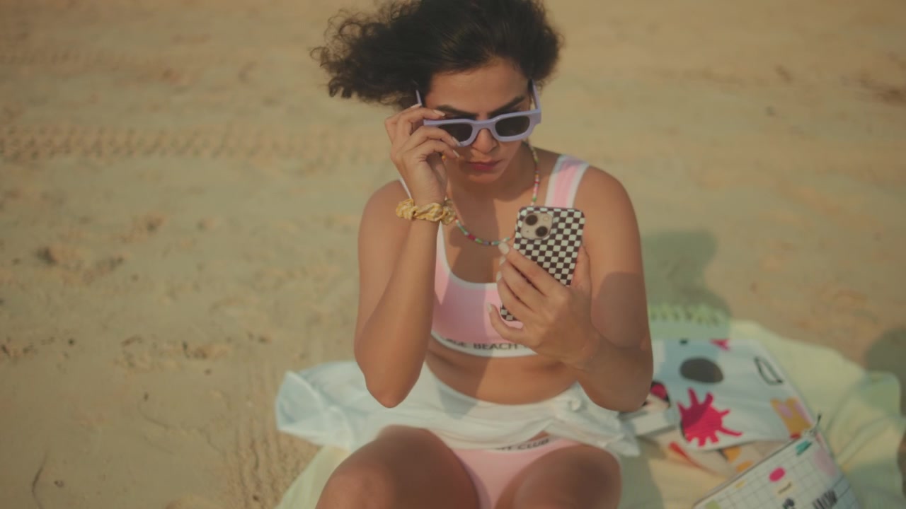 Donna in costume da bagno rosa seduta su una spiaggia sabbiosa che usa lo smartphone e si sistema gli occhiali da sole