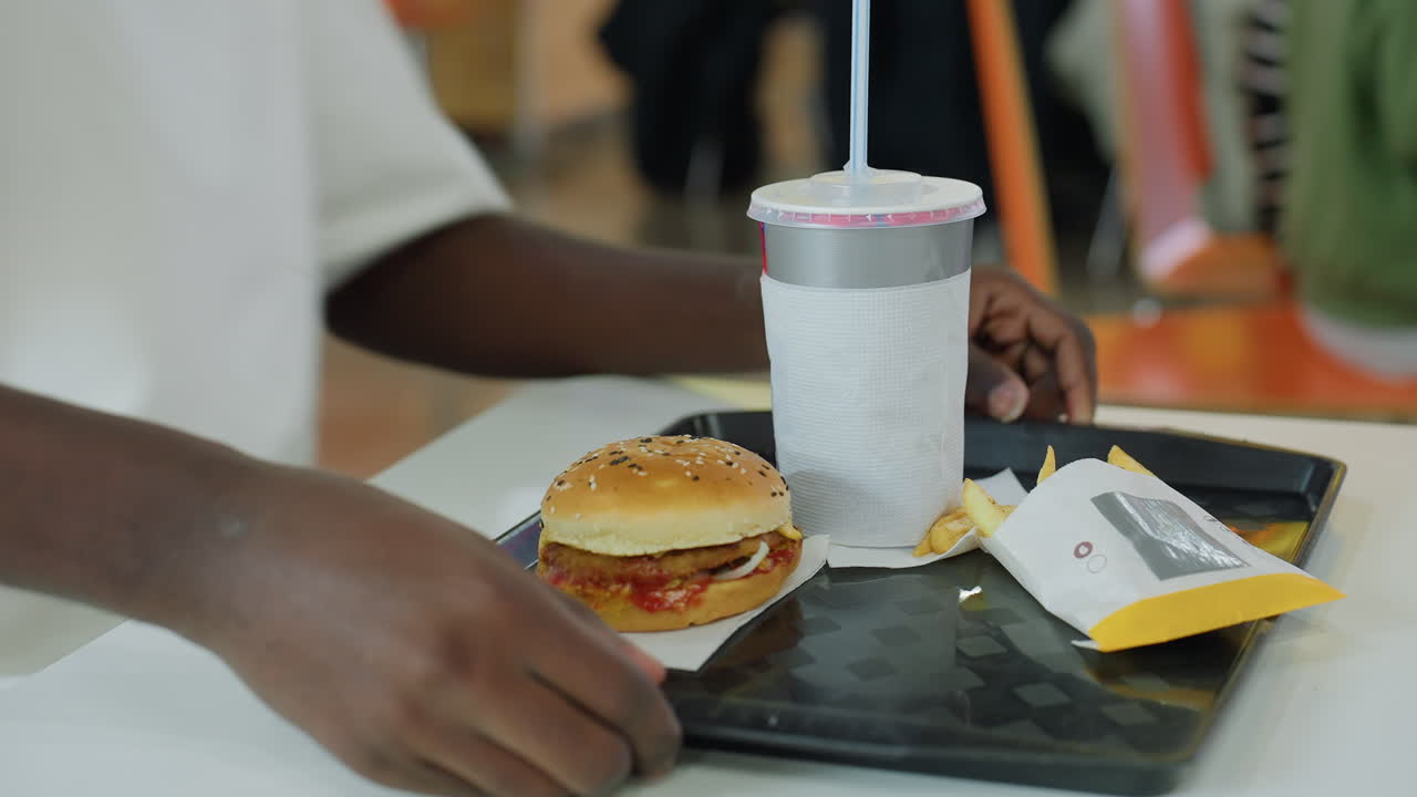 Young man wearing white shirt and jeans places tray with burger fries and soda cup on white table in bright casual cafe environment while preparing to eat quick meal surrounded by busy background