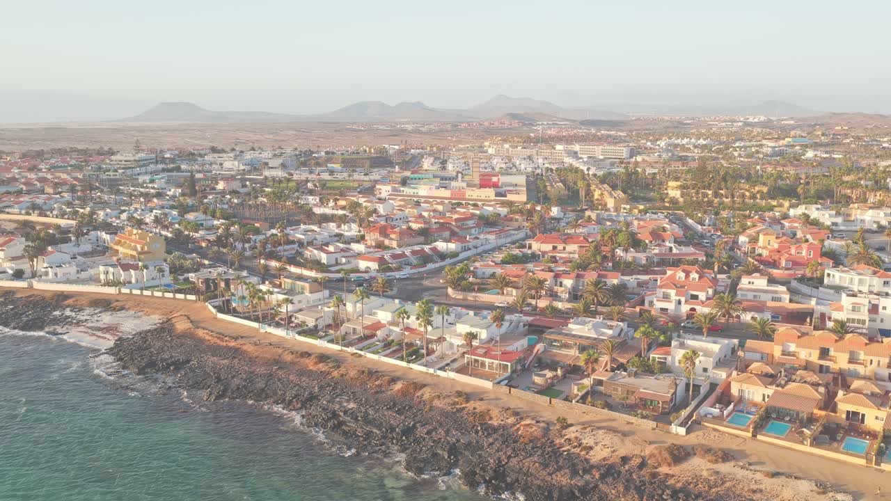 Warm sunlight highlighting pastel colored houses nestled along Playa del Medio waterfront in Fuerteventura, revealing charming Canary Islands coastal landscape, drone establishing shot