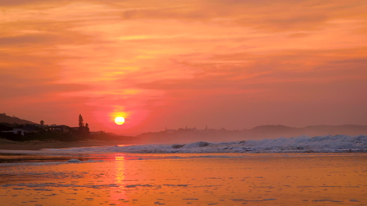 Vibrant orb of sun rises over beach, gold sky with low waves crashing, static shot