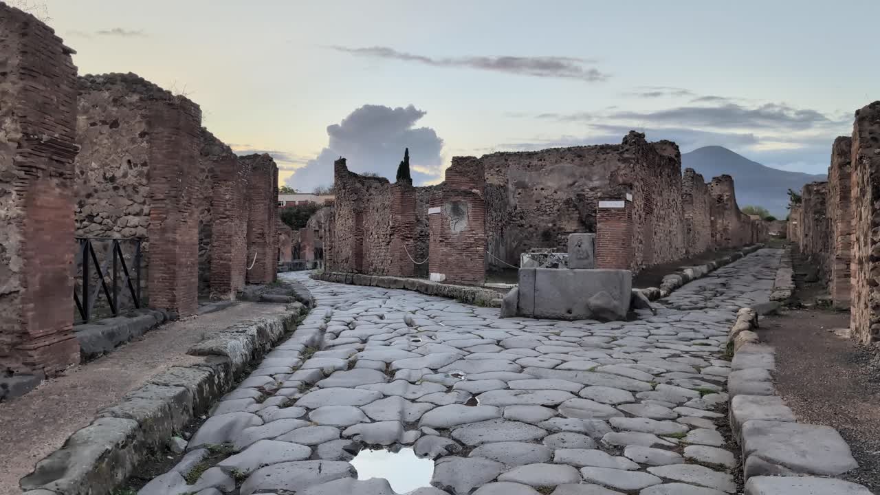 Slow pan of Pompeii’s ancient stone road glowing in golden sunset light, lined with ruins and framed by Mount Vesuvius in the distance. Perfect for travel or history footage