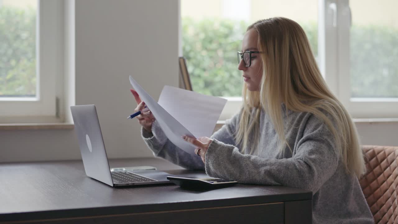 Una giovane donna con gli occhiali seduta a una scrivania, che esamina documenti mentre lavora al suo portatile a casa.