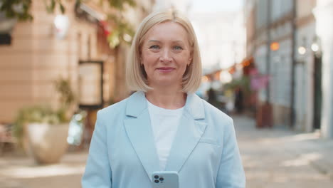 smiling mature caucasian businesswoman in suit using smartphone and looking at camera on city street