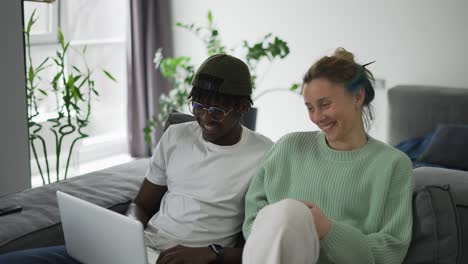 happy biracial couple sitting on sofa with laptop and talking