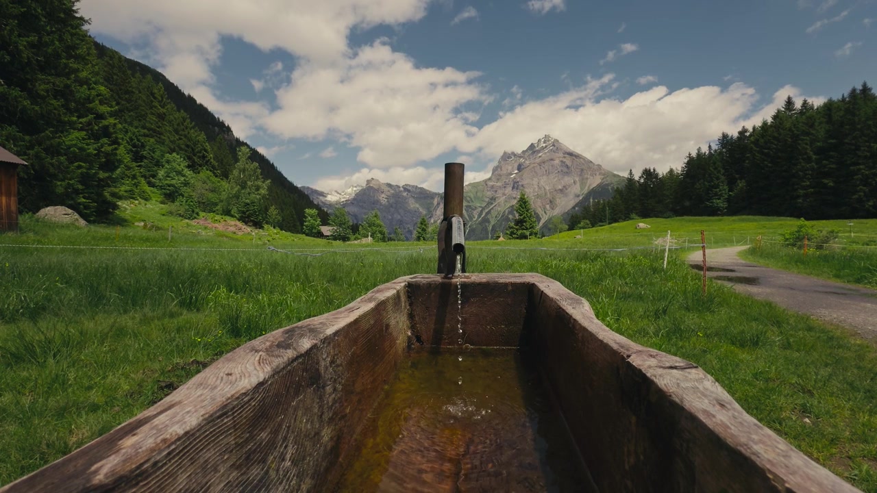 Fontaine à eau suisse traditionnelle avec de l'eau de montagne cristalline s'écoulant dans un bassin en bois. Des sommets alpins et un sentier de randonnée créent un paysage montagnard suisse authentique.