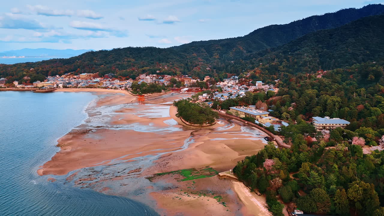 Iconic red Grand Torii Gate standing at the sandy beach of the Inland Sea at the Miyajima island, Japan. Picturesque seaside with beautiful shore, lovely private houses and stunning mountains. Aerial view.