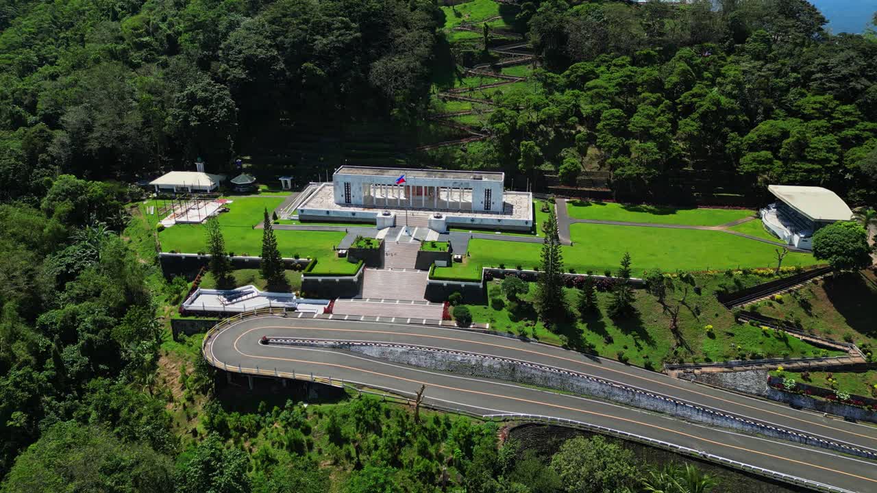 Tilt‑down aerial revealing the museum at the base of Mt. Samat National Shrine, framed by manicured lawns, pathways, and lush forested hills in Pilar, Bataan, Philippines
