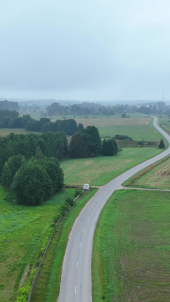 Drone shot flying above a winding countryside road, moving forward along fields, trees, and mist in the distance