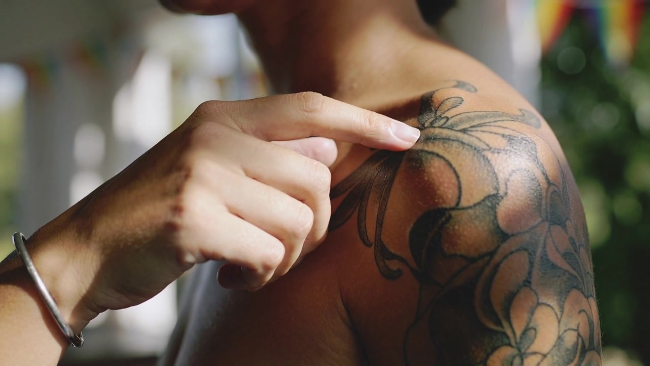 Una mano che preme, tracciando un tatuaggio floreale su una spalla nuda, percependo l'inchiostro, con un braccialetto da portico.