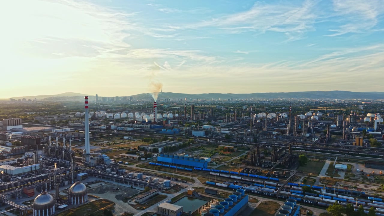 Et stort industriområde med fabrikker, skorsteiner og lagringsanlegg under solnedgang i et bylandskap.
