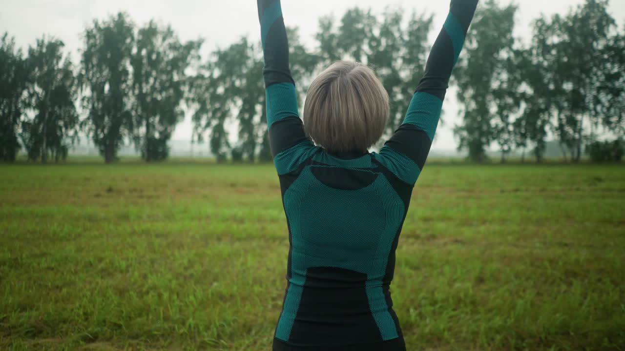 vista trasera de una mujer de cabello gris de pie con la cabeza ligeramente levantada, levantando los brazos en un movimiento sereno, practicando yoga en un vasto campo abierto bajo un cielo nublado con árboles borrosos en la distancia