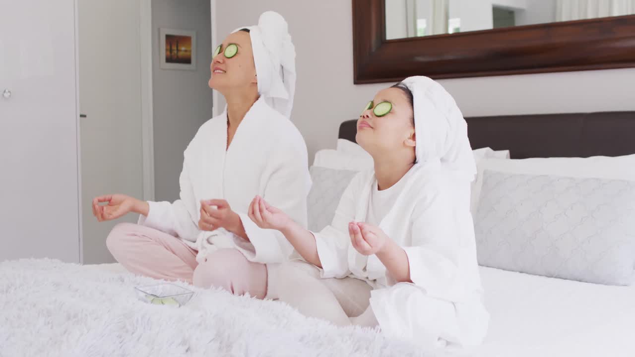 Video of happy asian mother and daughter in robes doing cucumber mask and resting