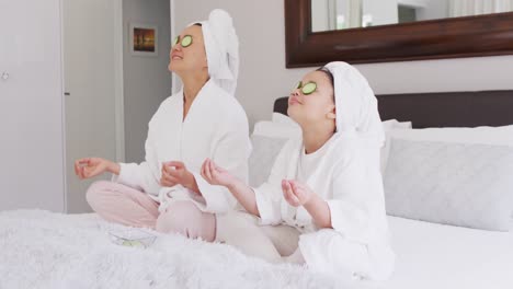 video of happy asian mother and daughter in robes doing cucumber mask and resting