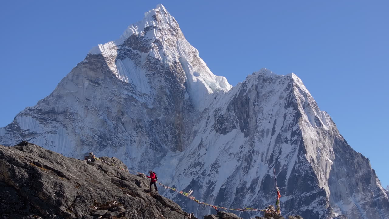 Un escursionista si dirige verso il Nangkartshang Peak, vicino a Dingboche, sulla rotta del Campo Base dell'Everest in Nepal. La maestosa Ama Dablam si erge sullo sfondo, mentre le bandiere di preghiera sventolano nel vento himalayano.