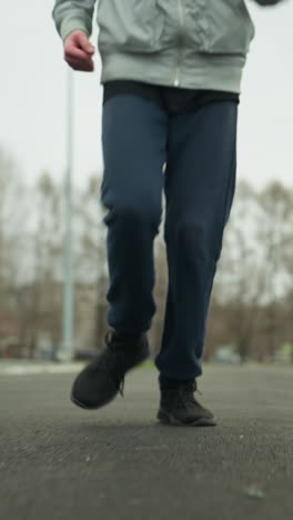 close-up leg view of two people jogging on a track in a stadium, the background shows blurred light poles and parked cars with bare trees