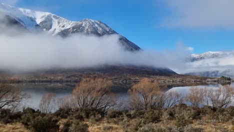 a-pan-from-right-to-left-of-a-small-lake-surrounded-by-snow-capped-mountains-with-low-fog-located-on-the-drive-to-Arthurs-Pass-in-Canterbury-New-Zealand