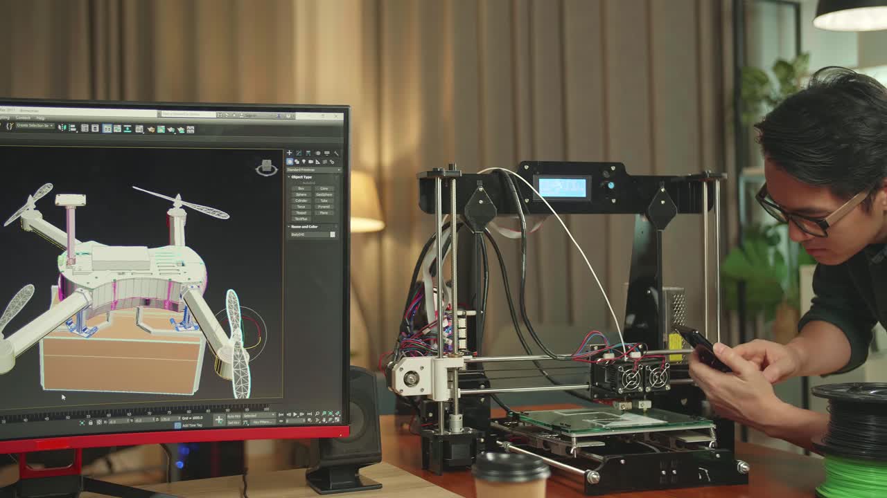 Asian Engineer Using Mobile Phone While Work On 3D Printer Printing And Personal Computer, Screen Shows Cad Software With 3D Prototype Of Drone Delivery