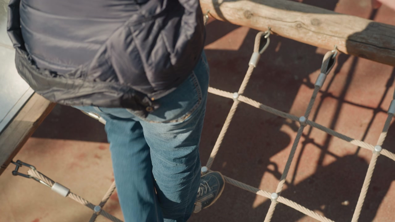Primo piano delle gambe e dei piedi di una persona che attraversa con cautela un ponte di corda all'aperto, in equilibrio su corde spesse con connettori metallici. La persona indossa jeans blu e scarpe da ginnastica scure mentre si muove.