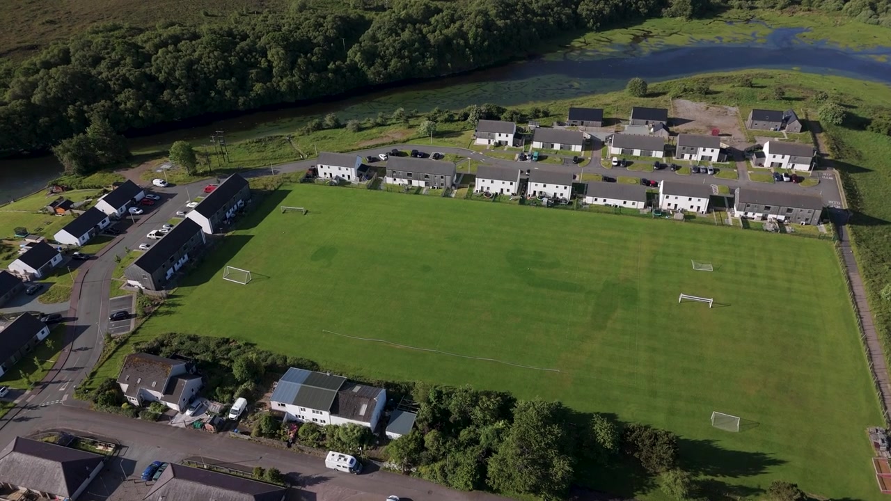 Cinematic aerial shot of a lush green football pitch in a residential neighborhood by a river, showcasing community wellness and ecofriendly living.