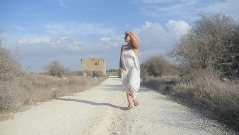 wide shot of a woman in a white dress searches for something down a deserted dirt path with ruins in the background