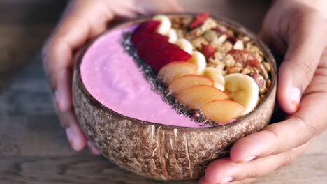 cerca de las manos de una mujer sosteniendo un tazón de batido rosa con frutas, granola y semillas de chía