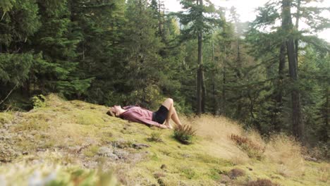 a beautiful young adult woman lies down on green and mossy forest ground while enjoying sunshine and calmness