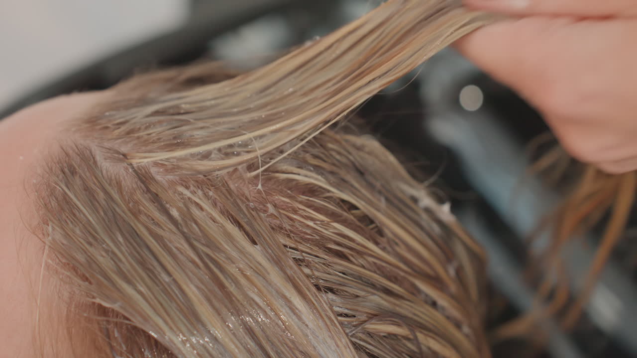 Application d’une coloration blonde dans un lavabo de salon, gros plan sur les mains d’une coiffeuse appliquant un toner crémeux sur des mèches mouillées tandis que la cliente est allongée au lavabo. Des ongles noirs vernis maintiennent les cheveux sectionnés tandis qu’une brosse étale le produit.