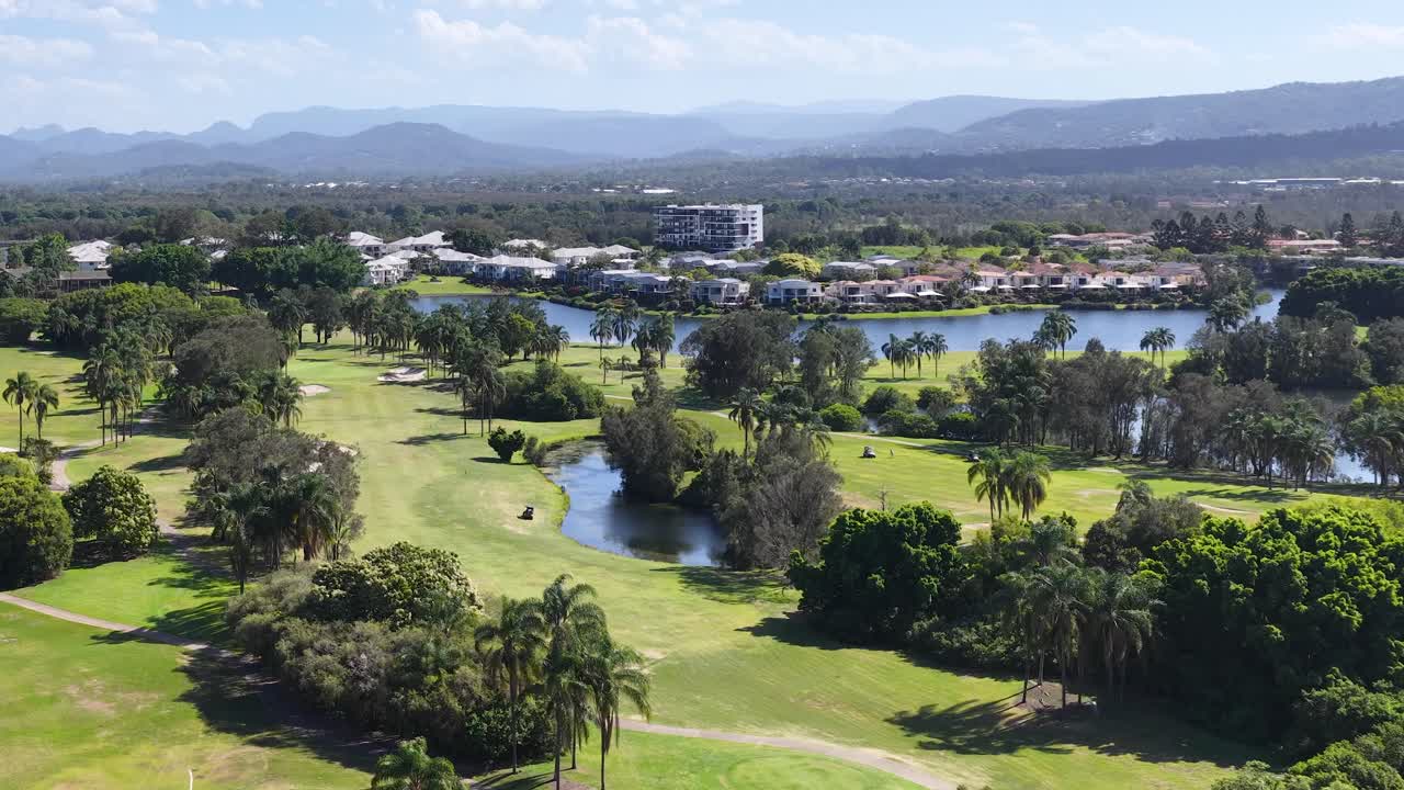 Sunny aerial drone pan across lush golf course, winding lake, residential waterfront, and distant mountains