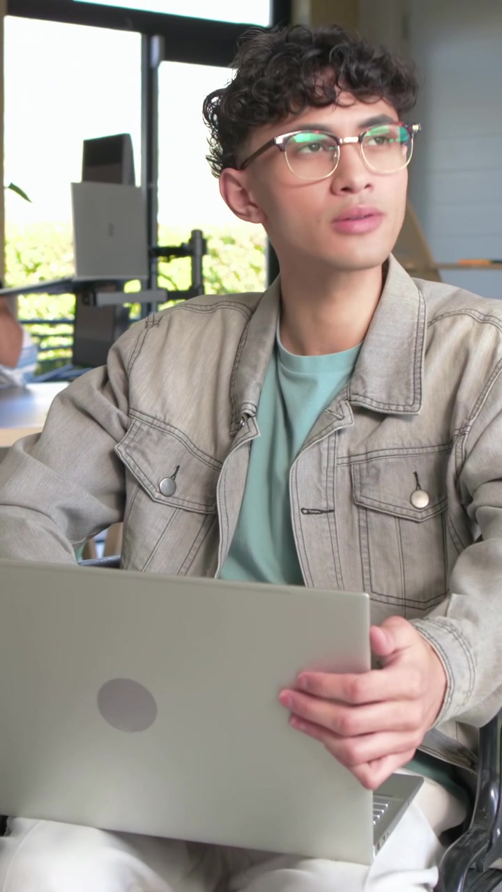 Vertical video: Typing adult man working with laptop on lap at home, adjusting glasses, wheelchair